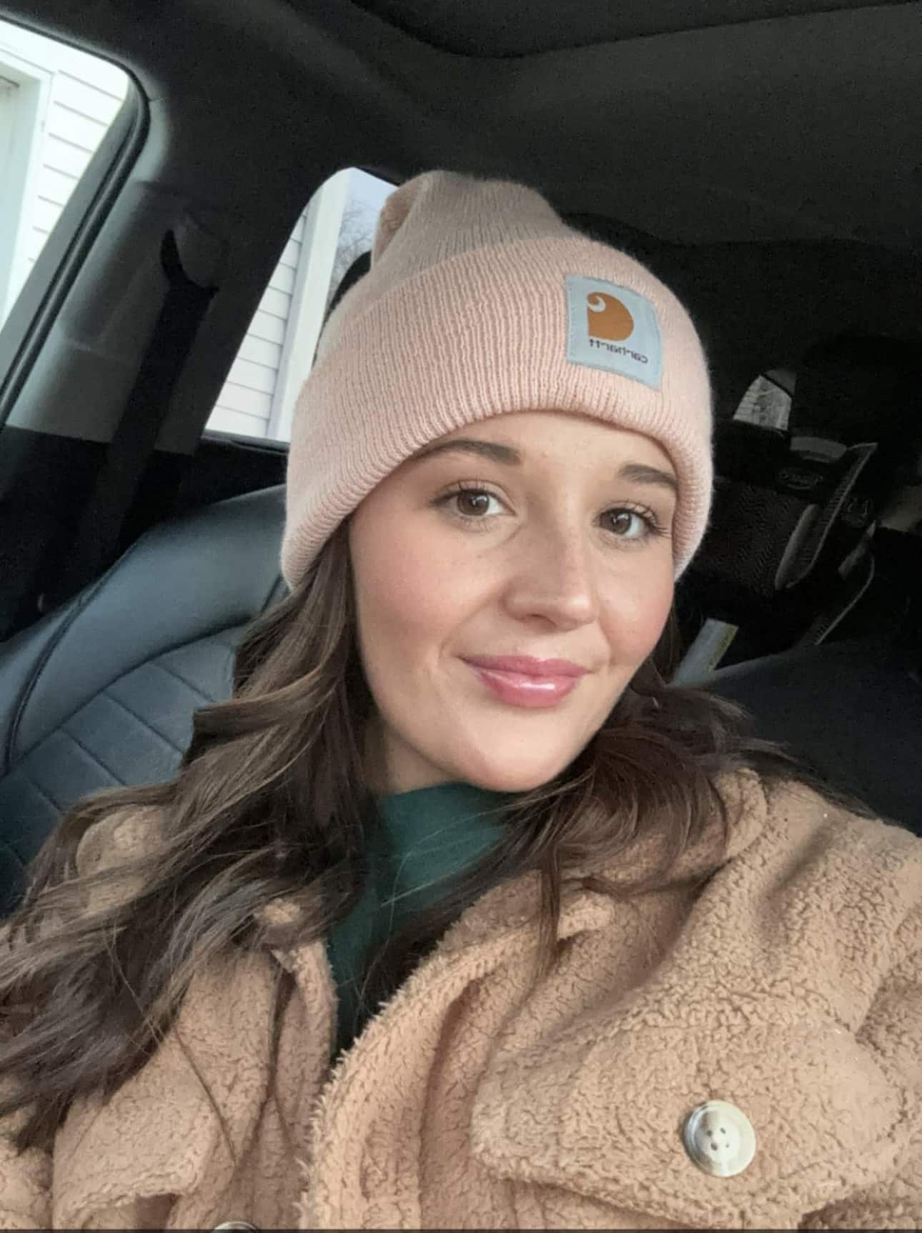 A woman with long brown hair sits in a car, wearing a pink beanie and a tan fleece jacket. She is smiling softly and looking at the camera. The car interior and part of a window are visible in the background.