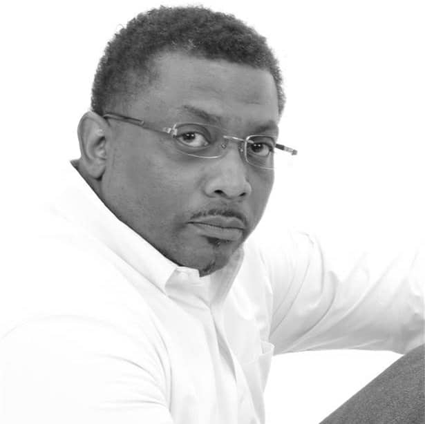 Black-and-white photo of a man with short, curly hair and glasses, wearing a light-colored button-up shirt, looking directly at the camera with a neutral expression.