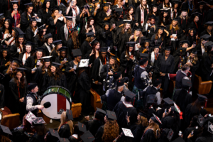 A large group of graduates in black caps and gowns stand together, many smiling and taking photos. A person with a decorated drum leads a procession through the crowd. The atmosphere is festive and celebratory.