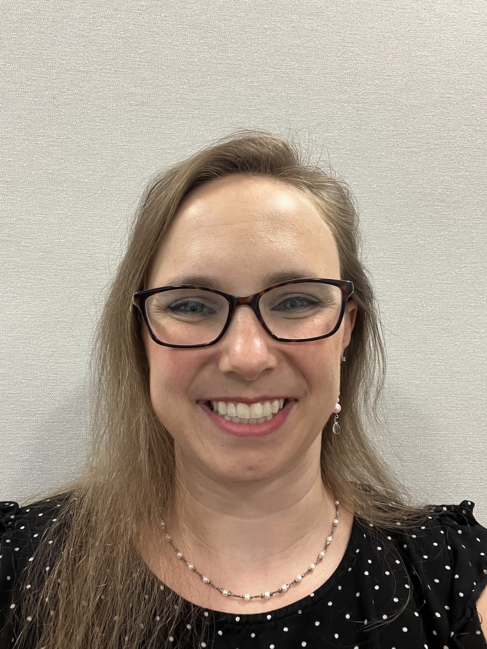 A woman with long light brown hair, wearing glasses and a black polka dot top, smiles at the camera. She has on a beaded necklace and dangling earrings, with a light-colored textured background behind her.