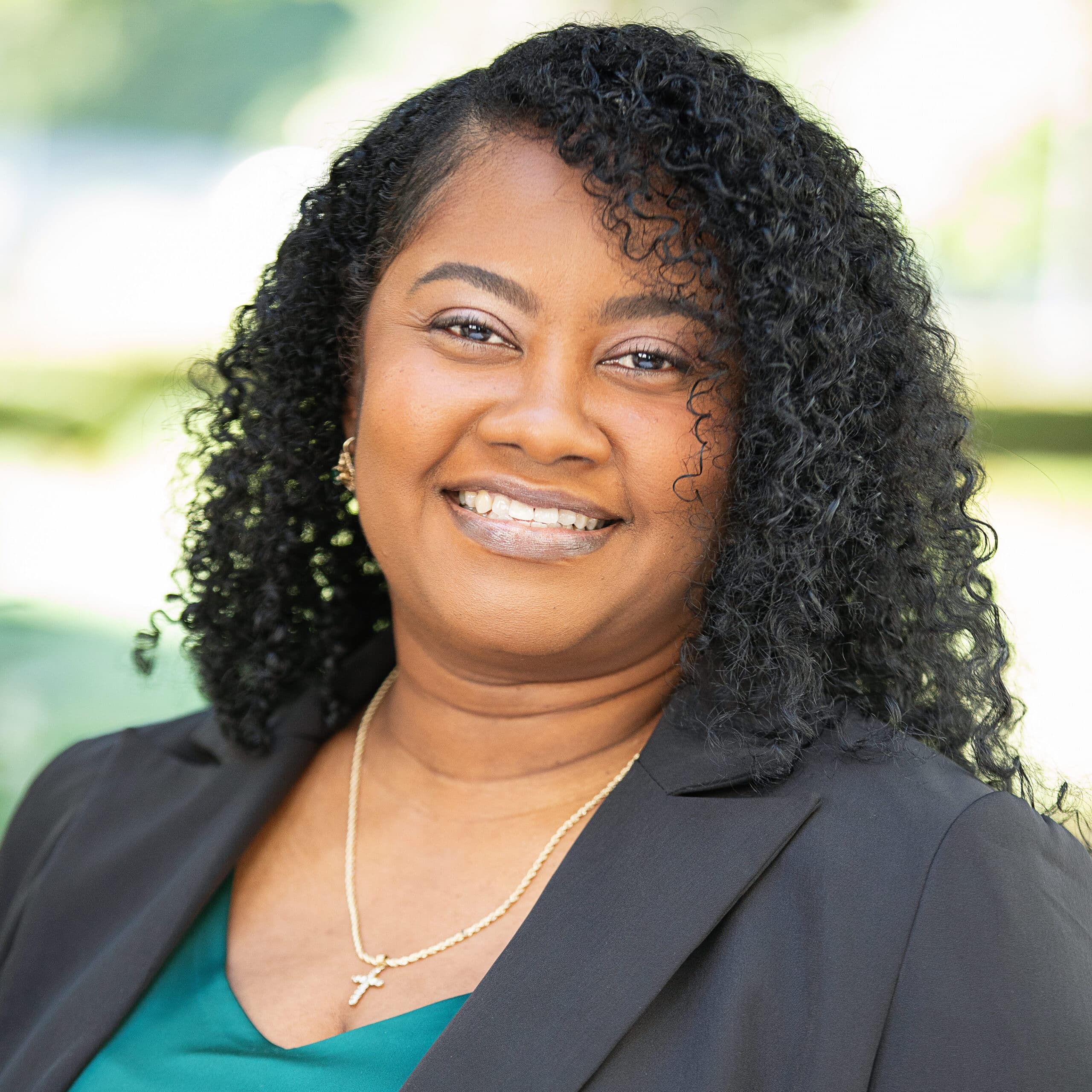 A woman with curly hair smiles while wearing a black blazer over a teal top. She has a gold necklace with a cross pendant and is standing against a blurred outdoor background.