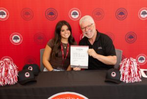 A smiling young woman and an older man sit at a table with red pom-poms and hats, holding up a certificate. A red backdrop with circular logos is behind them.