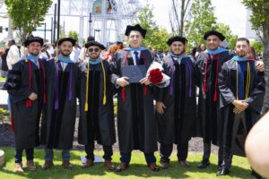 Seven men in graduation gowns and caps stand outdoors, smiling for a group photo. One holds a diploma and bouquet of roses. Trees and a crowd are visible in the background on a sunny day.