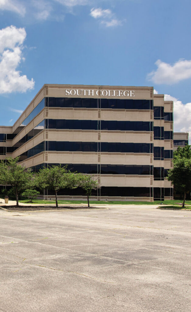 A modern, multi-story building with large glass windows displays the sign "South College" on top. In front, there are small trees and an empty parking lot under a partly cloudy sky.