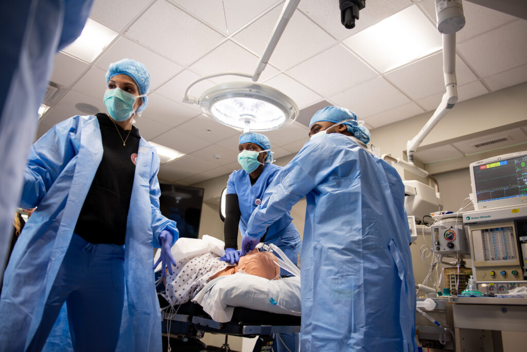 Four medical professionals in masks and gowns treat a patient on a hospital bed in a brightly lit room with medical equipment and monitors visible.