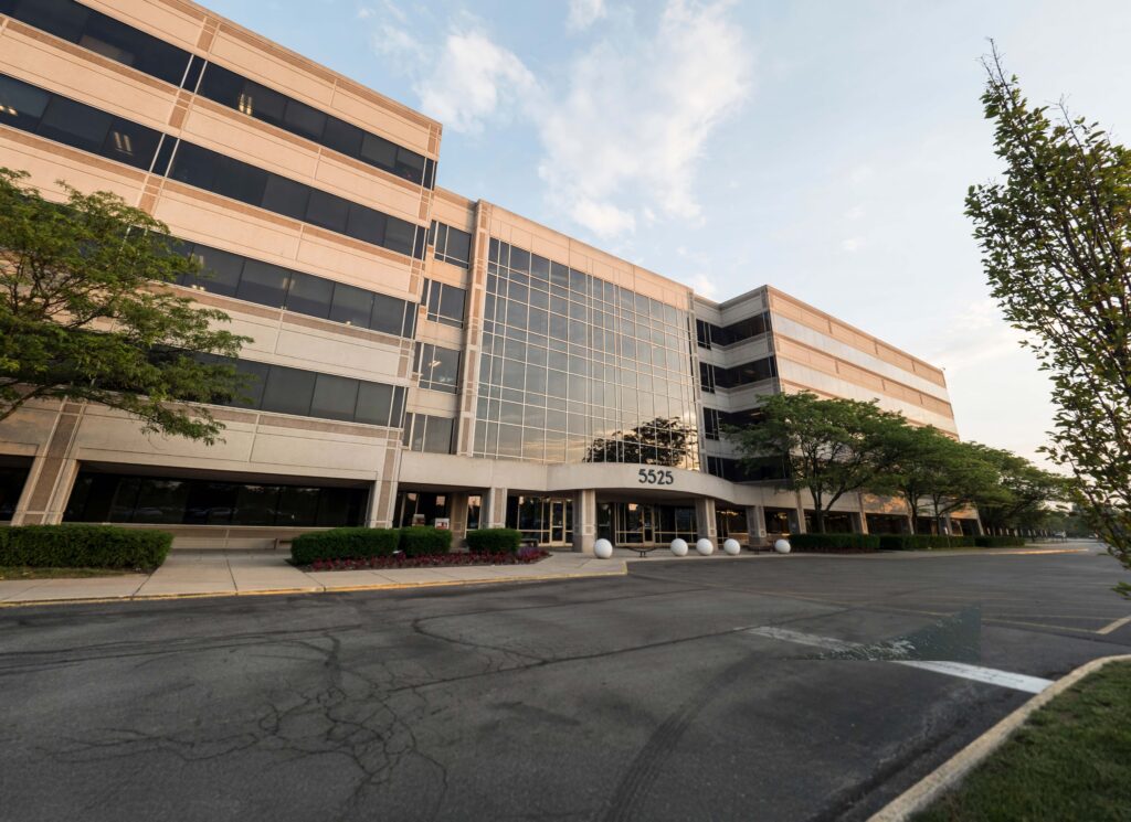A modern office building with large windows, reflective glass, and the number 5525 above the entrance. Trees and bushes line the walkway, and the parking lot is mostly empty.