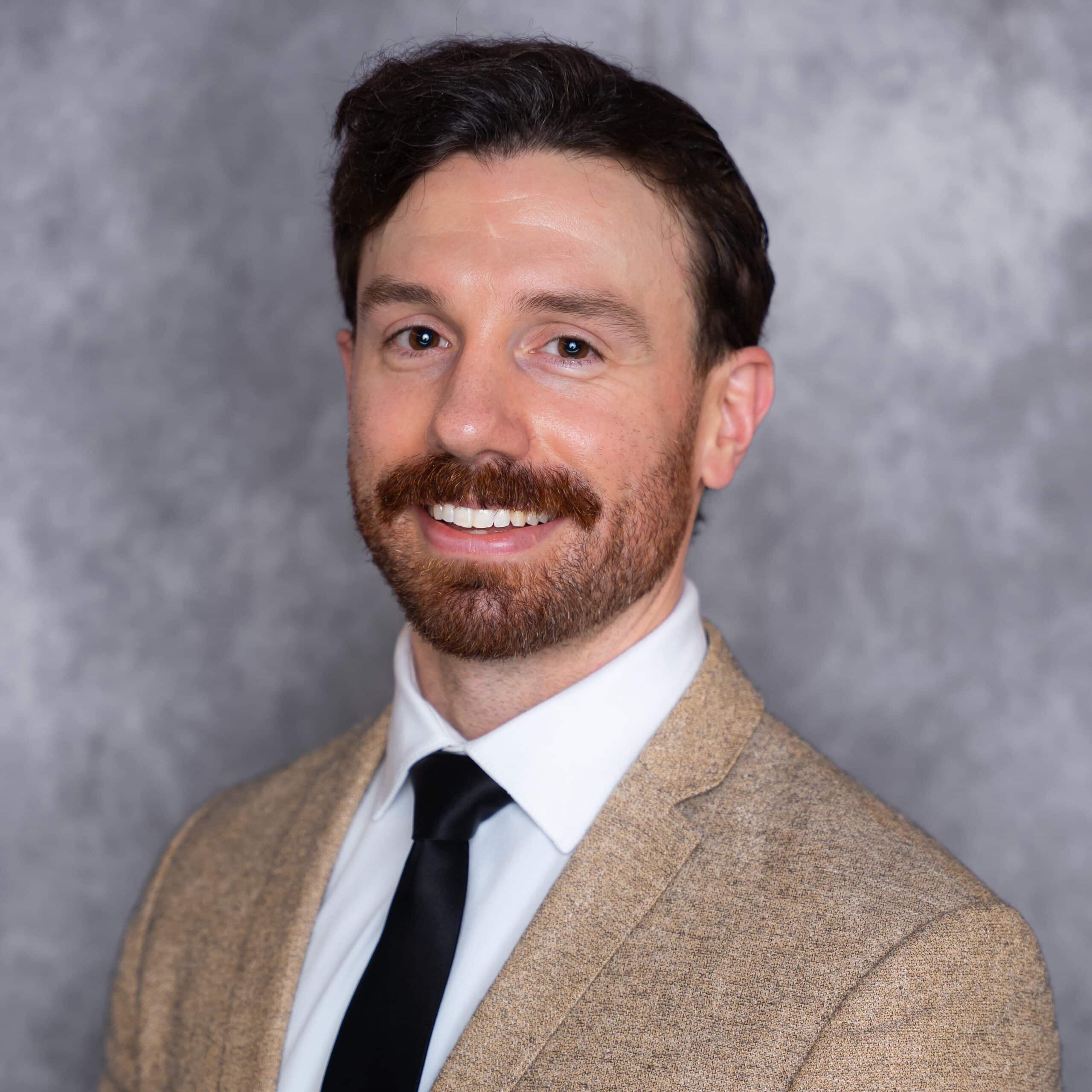A man with brown hair and a mustache, wearing a tan suit jacket, white shirt, and black tie, smiles at the camera against a gray mottled background.