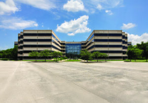 A wide, modern building labeled "South College" with glass entrance, striped facade, symmetrical wings, and an empty parking lot under a blue sky with scattered clouds.