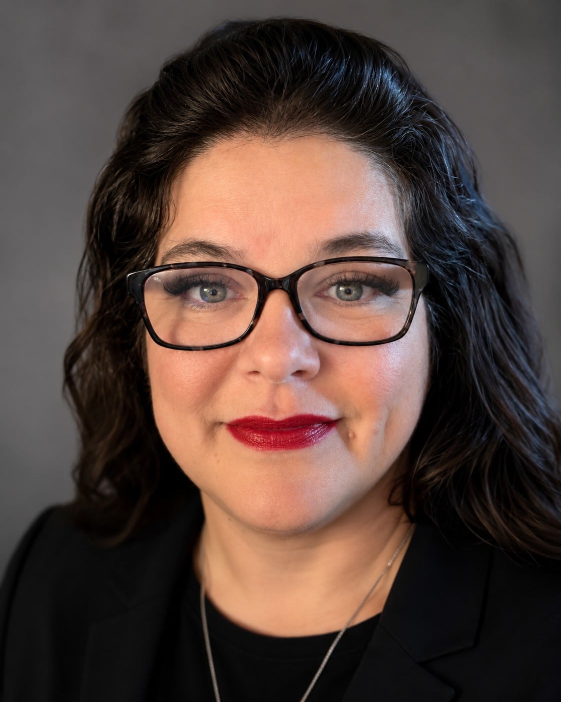 Woman with long dark wavy hair, wearing glasses, a black blazer, and red lipstick, smiling slightly in front of a neutral gray background.