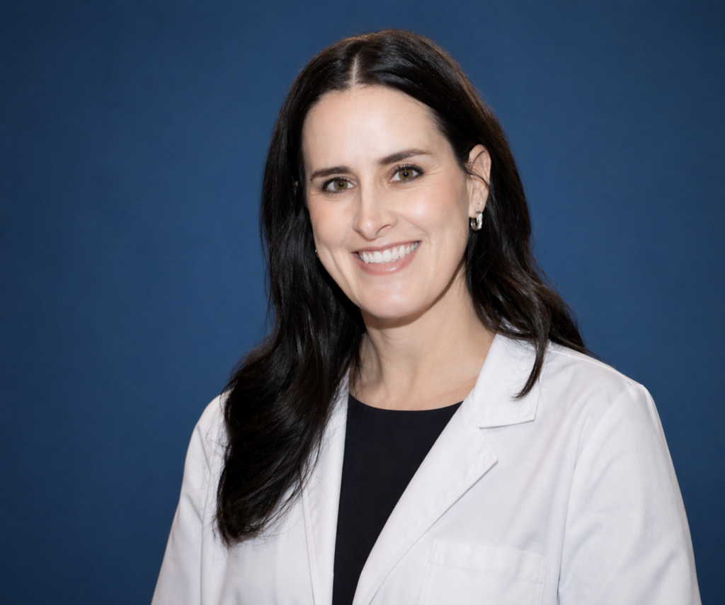 Woman with long dark hair wearing a white lab coat and black shirt, smiling in front of a solid blue background.