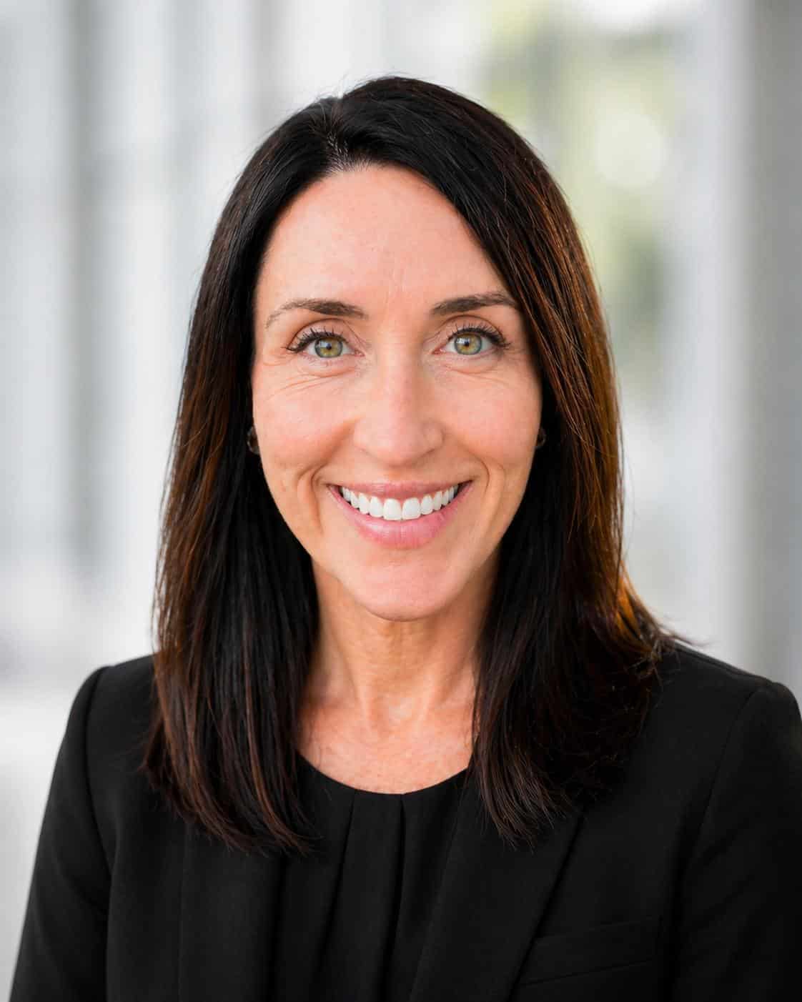 A woman with straight brown hair and green eyes smiles at the camera. She is wearing a black blazer and a black top. The background is softly blurred with natural light.