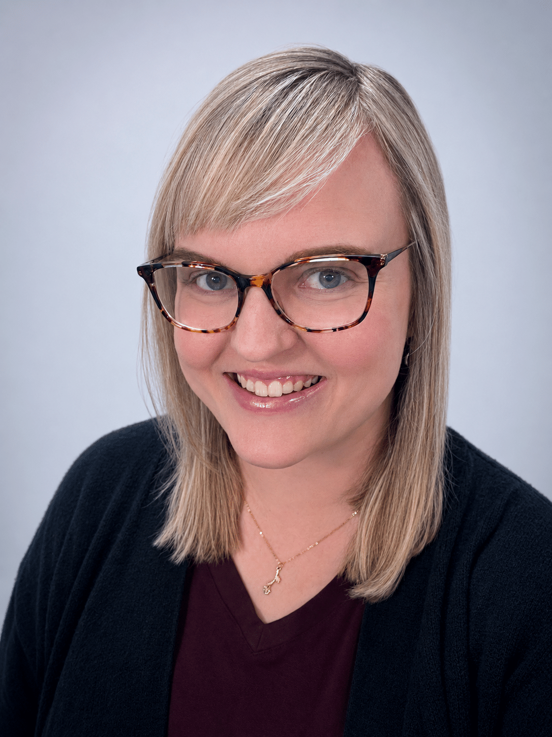 A woman with straight blonde hair, wearing tortoiseshell glasses, a dark top, and a black cardigan, smiles at the camera against a plain light background.