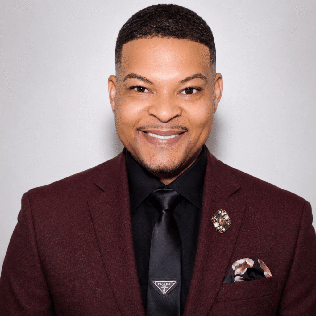 A man in a burgundy suit, black shirt, and black Prada tie smiles at the camera. He has short hair, a brooch and pocket square on his jacket, and stands against a plain light background.