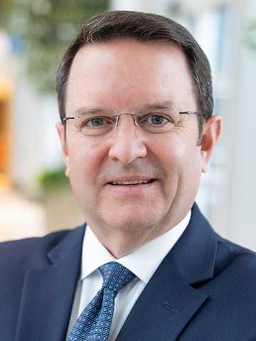A man wearing glasses, a dark suit, white shirt, and patterned blue tie, smiling slightly in an indoor setting with blurred background.