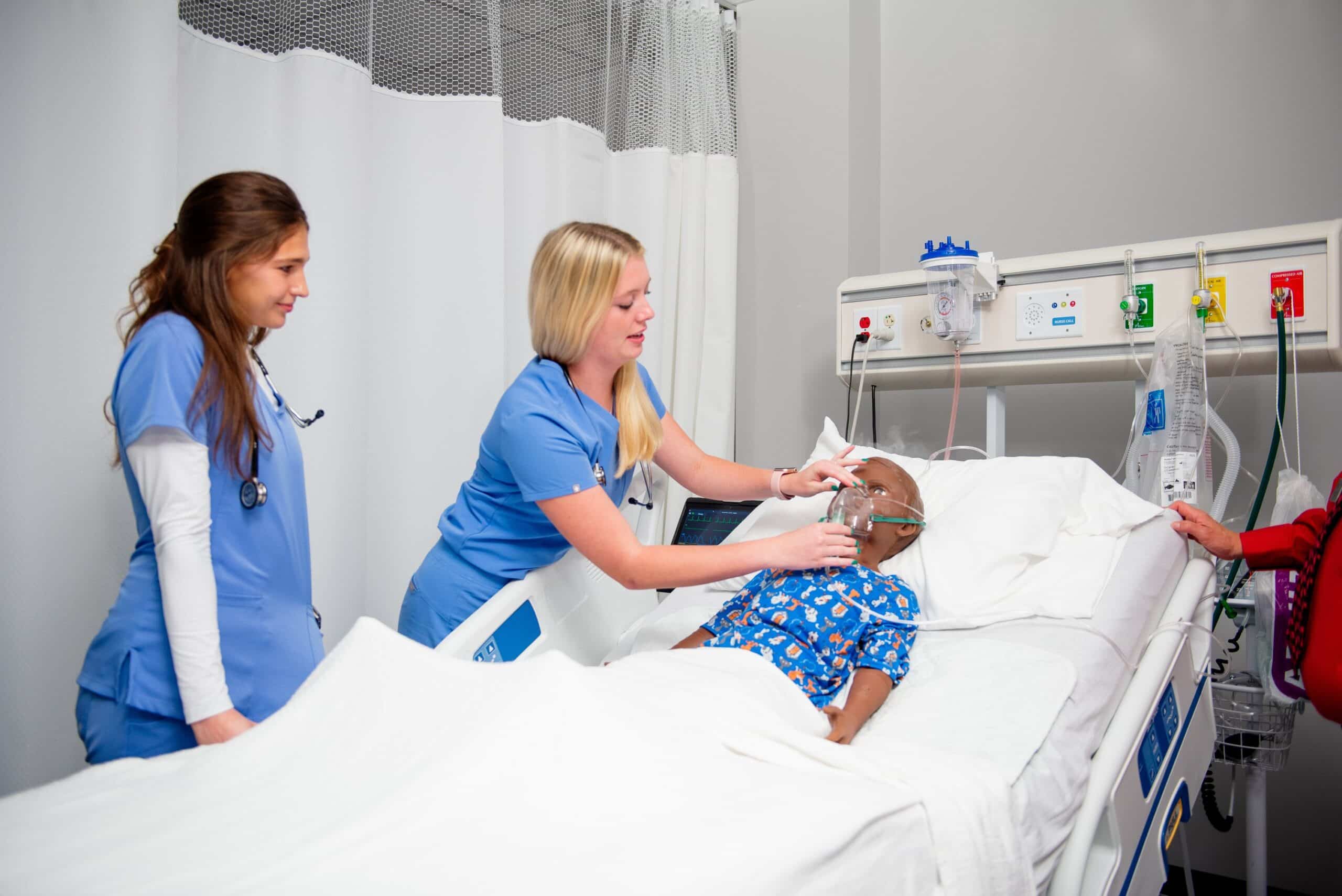 Two nurses in blue scrubs attend to a child in a hospital bed, adjusting an oxygen mask on the patient, surrounded by medical equipment and white curtains.