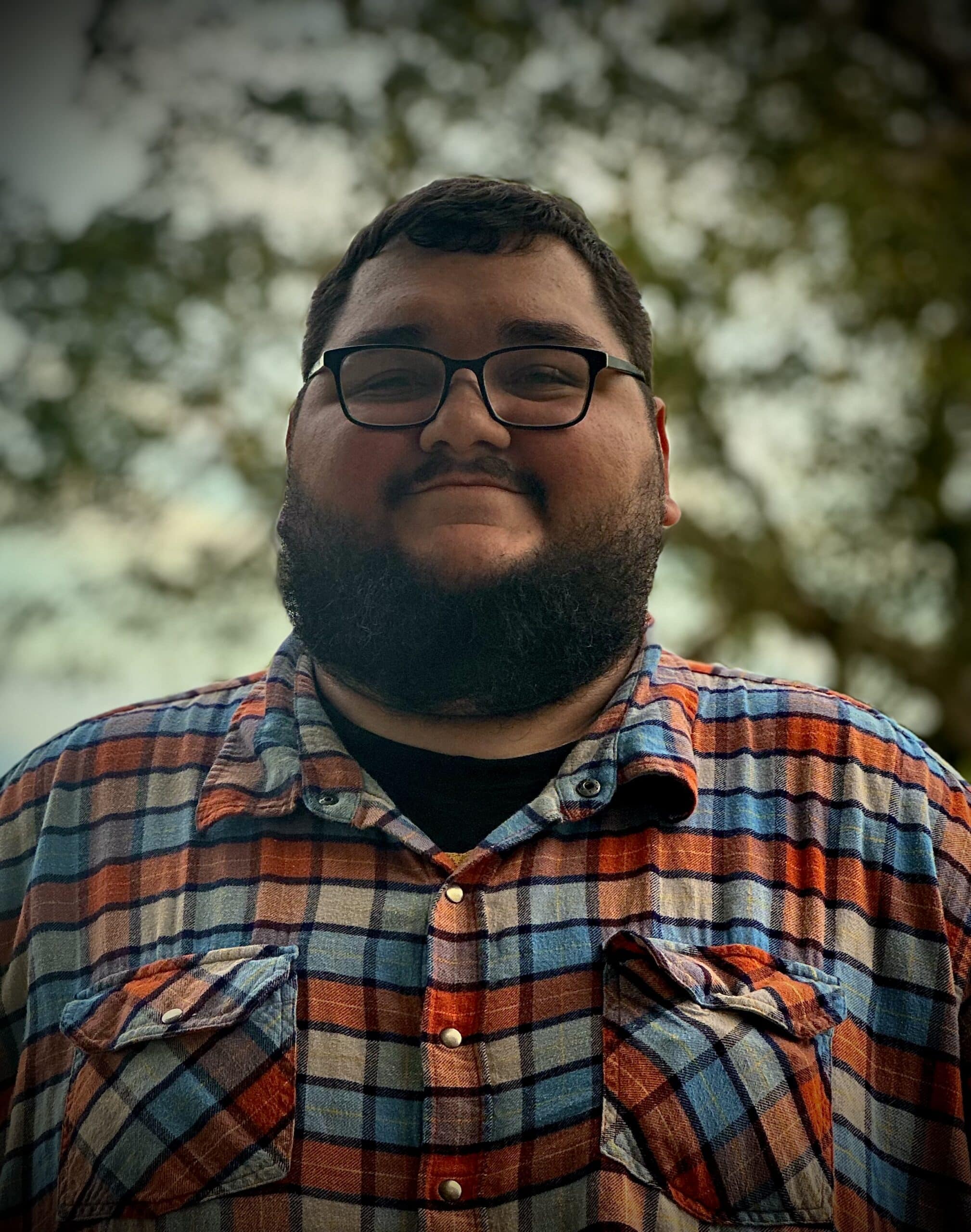 A man with a beard and glasses is smiling outside, wearing a plaid shirt with blue, orange, and white colors. Trees and a cloudy sky are visible in the background.