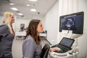 Two healthcare professionals in gray scrubs operate an ultrasound machine in a clinical room. One focuses on the monitor displaying an ultrasound image while the other stands nearby, smiling.
