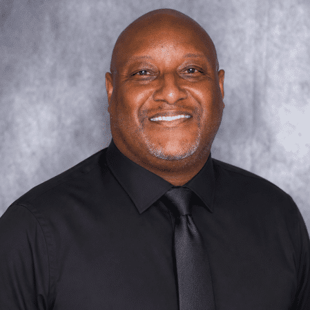 A man smiles at the camera, wearing a black dress shirt and black tie. The background is a plain, light gray surface.