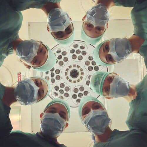 Eight surgeons in scrubs and masks stand in a circle, looking down towards the camera, with an operating room light fixture above them in the background.
