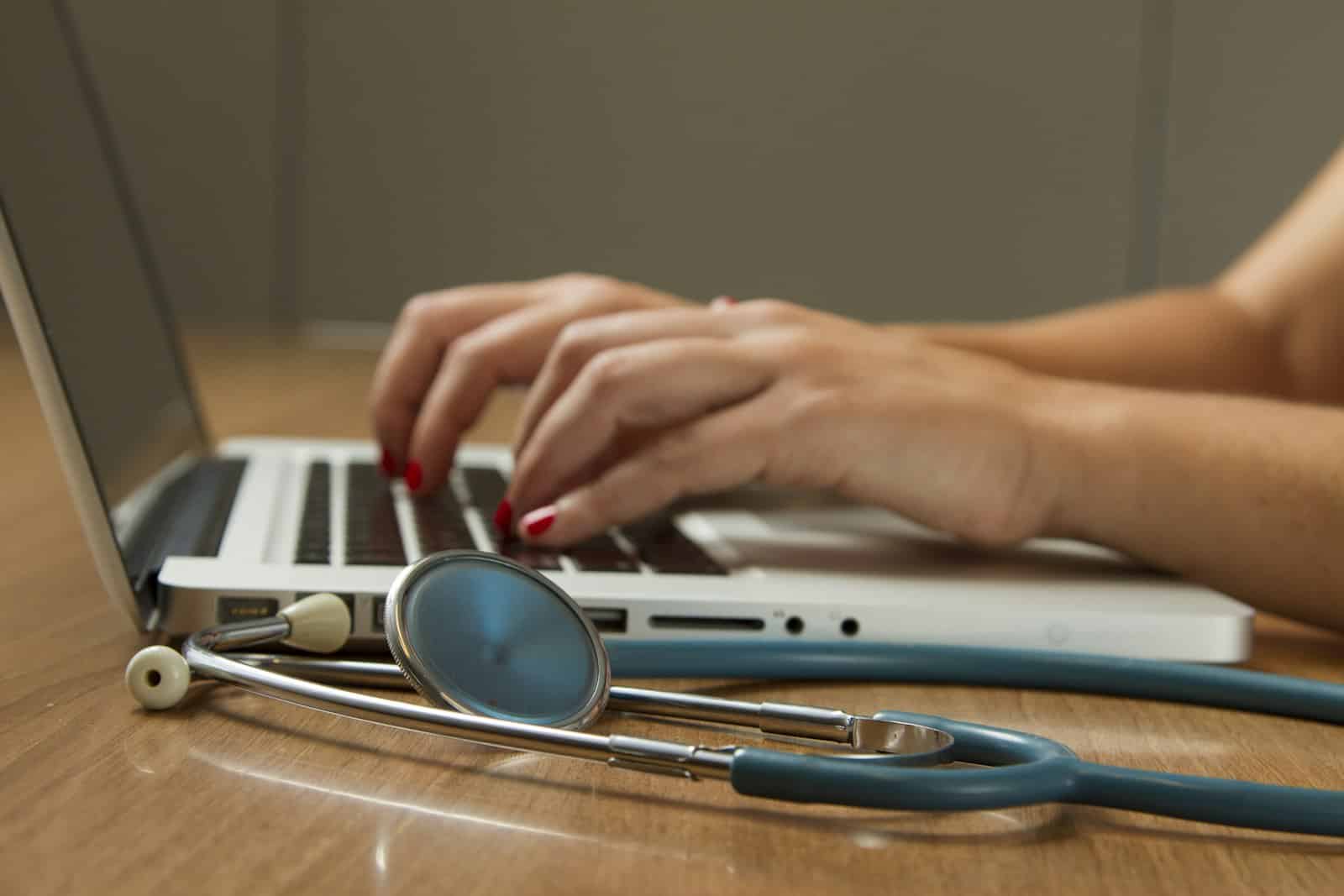 Hands typing on a laptop keyboard with a stethoscope lying on the wooden desk, suggesting a healthcare or medical professional working on a computer.