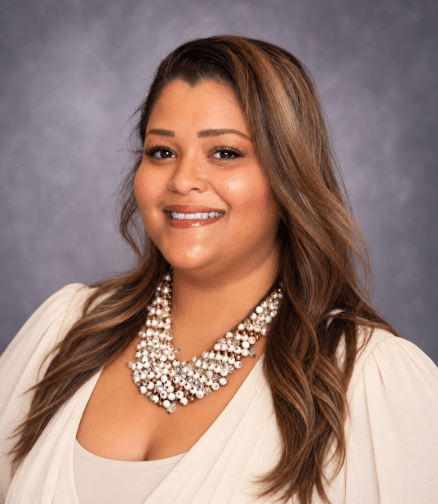 A woman with long brown hair smiles, wearing a cream-colored top and a chunky, multi-strand pearl necklace.